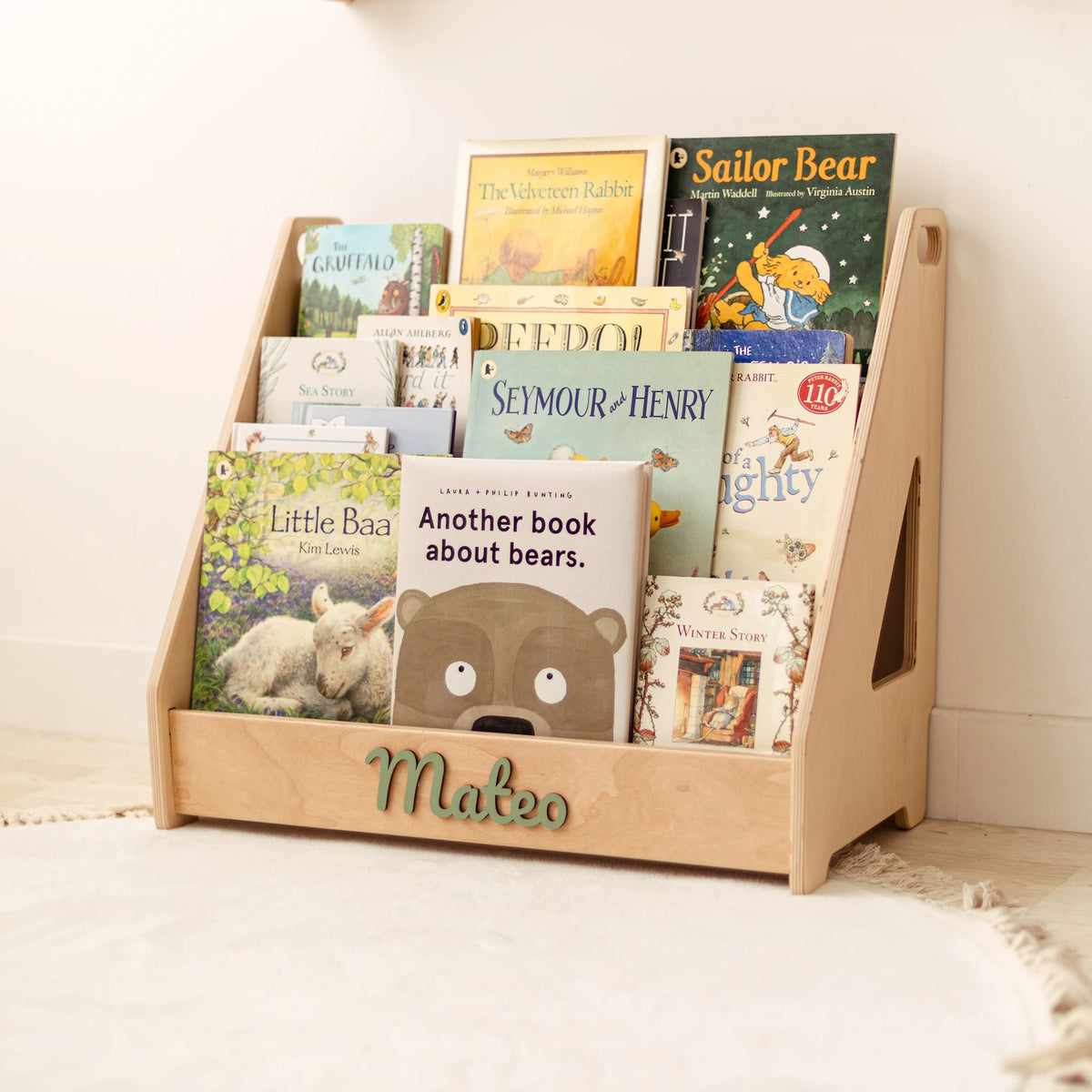 Light wood Montessori front-facing bookshelf with name Mateo in green letters, full of picture books in a bright nursery