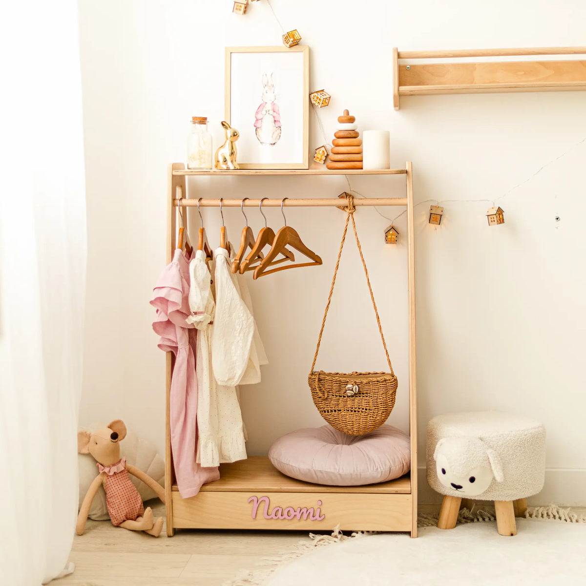 Light wood Montessori kids' clothing rack with hanging clothes and a basket, name Naomi in a styled corner