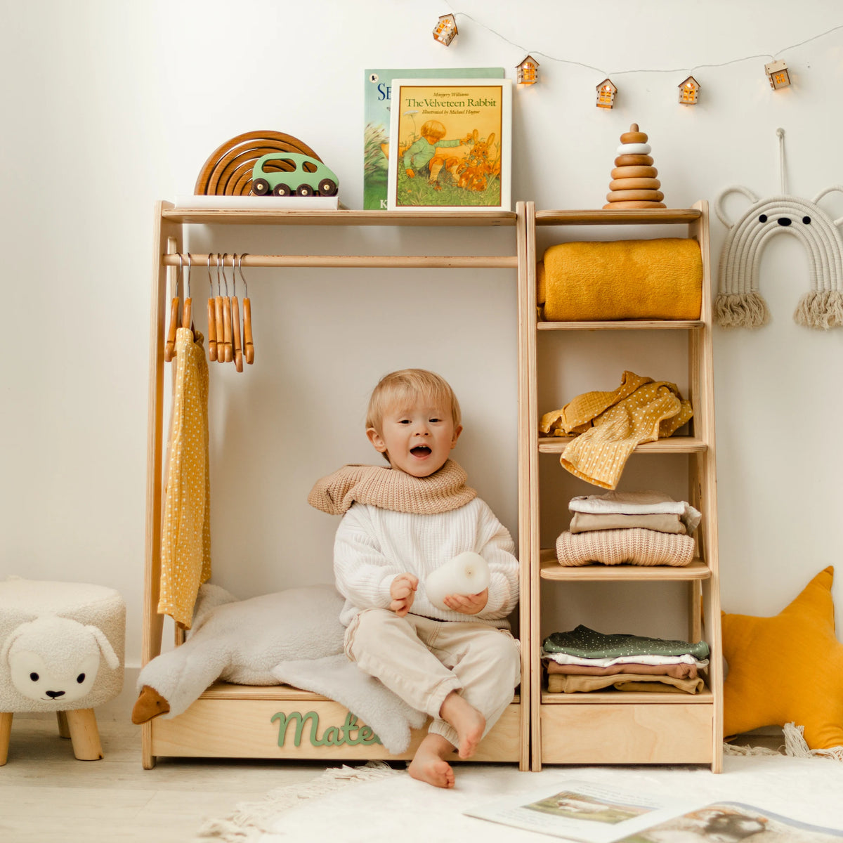 Light wood kids' clothing rack and shelf with a baby sitting on the base and folded clothes, name Mateo