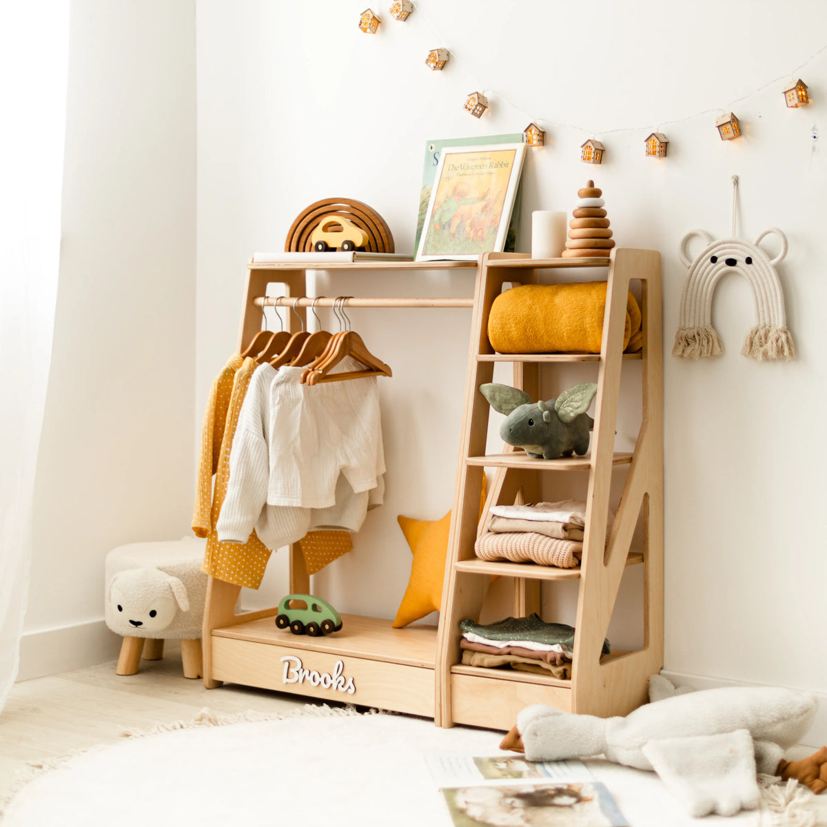 Light wood Montessori kids' clothing rack and open shelf set with hanging and folded clothes, name Brooks, in a nursery
