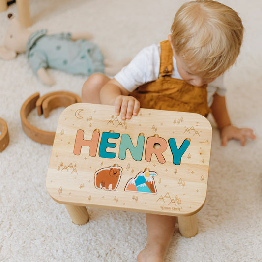 Little boy holding a personalized wooden name puzzle stool featuring the name Henry and printed Wildwood theme elements.
