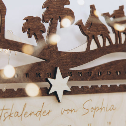 Wooden Nativity advent calendar with camels, palm trees and a star ornament, string lights in background